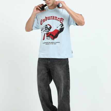 Man wearing a light blue t-shirt with graphic design and text, paired with dark jeans on a white background.